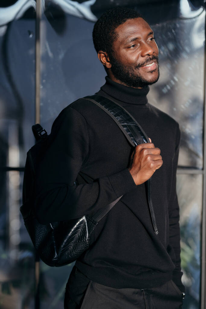 Αφροαμερικάνος με μαύρο παντελόνι και πουκάμισο κρατώντας μια τσάντα στην πλάτη του χαμογελώντας - Φωτογραφία, εικόνα
