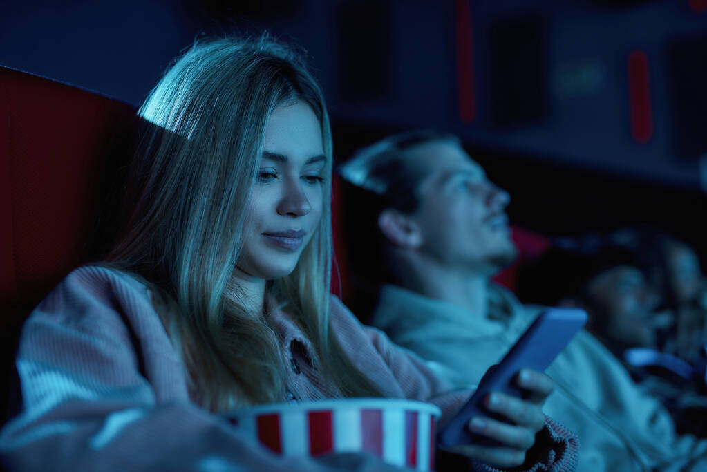 Portrait of beautiful young woman smiling, using her phone while missing boring movie at the cinema - Photo, Image