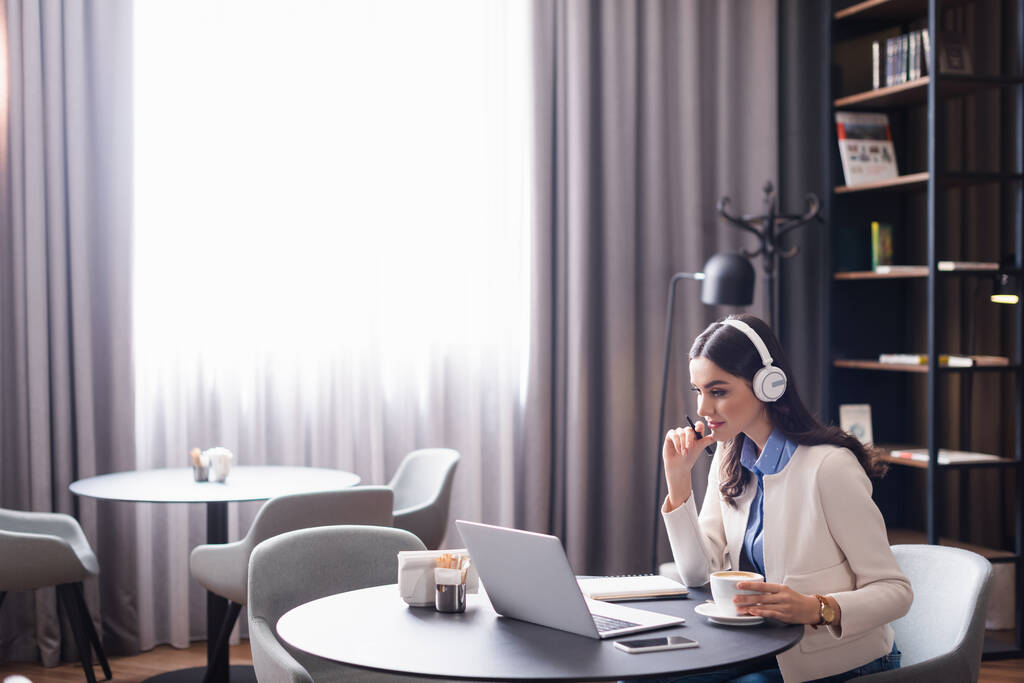 концентрированный фрилансер в наушниках, работающих рядом с ноутбуком и чашкой кофе в ресторане - Фото, изображение