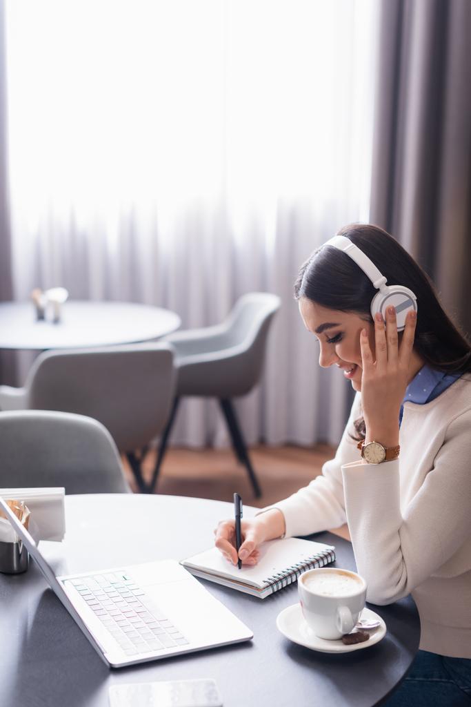 freelancer positivo tocando fones de ouvido sem fio enquanto escreve em notebook no restaurante - Foto, Imagem