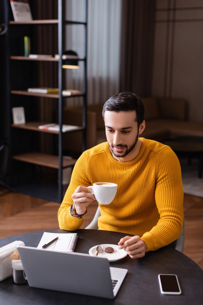 fiatal arab szabadúszó kezében csésze kávét, miközben nézi laptop étteremben - Fotó, kép