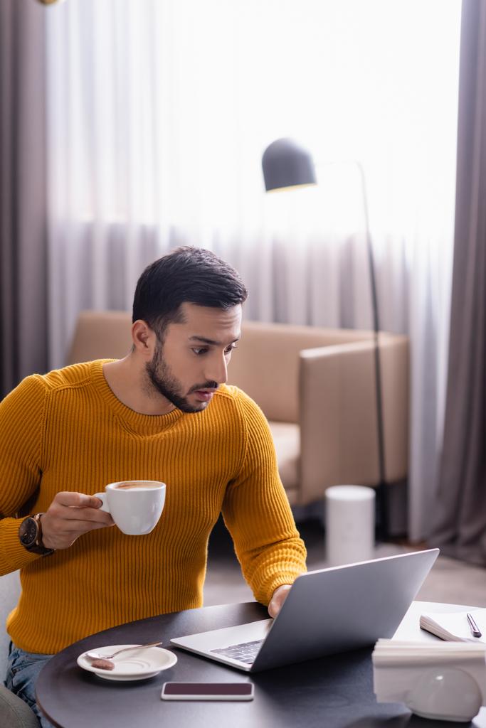 freelance arabo sorpreso che tiene la tazza di caffè mentre guarda il computer portatile nel ristorante - Foto, immagini