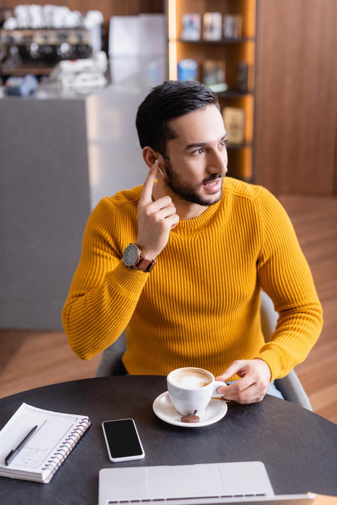 joven árabe freelancer fijación de auriculares mientras está sentado cerca de la taza de café y portátil - Foto, imagen