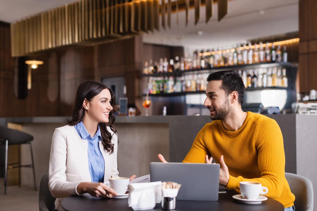 jonge Arabische freelancer praten met vrolijke vriend in de buurt van laptop en koffiekopjes in restaurant, wazig voorgrond - Foto, afbeelding