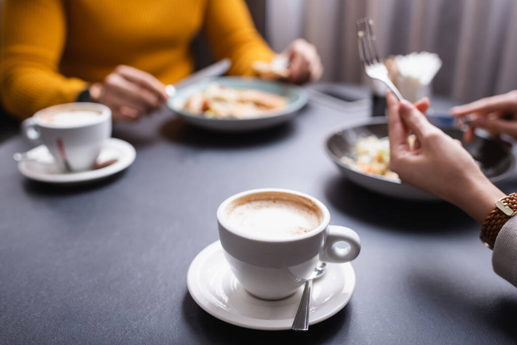 Vista recortada de cappuccino cerca de pareja cenando en restaurante sobre fondo borroso  - Foto, imagen