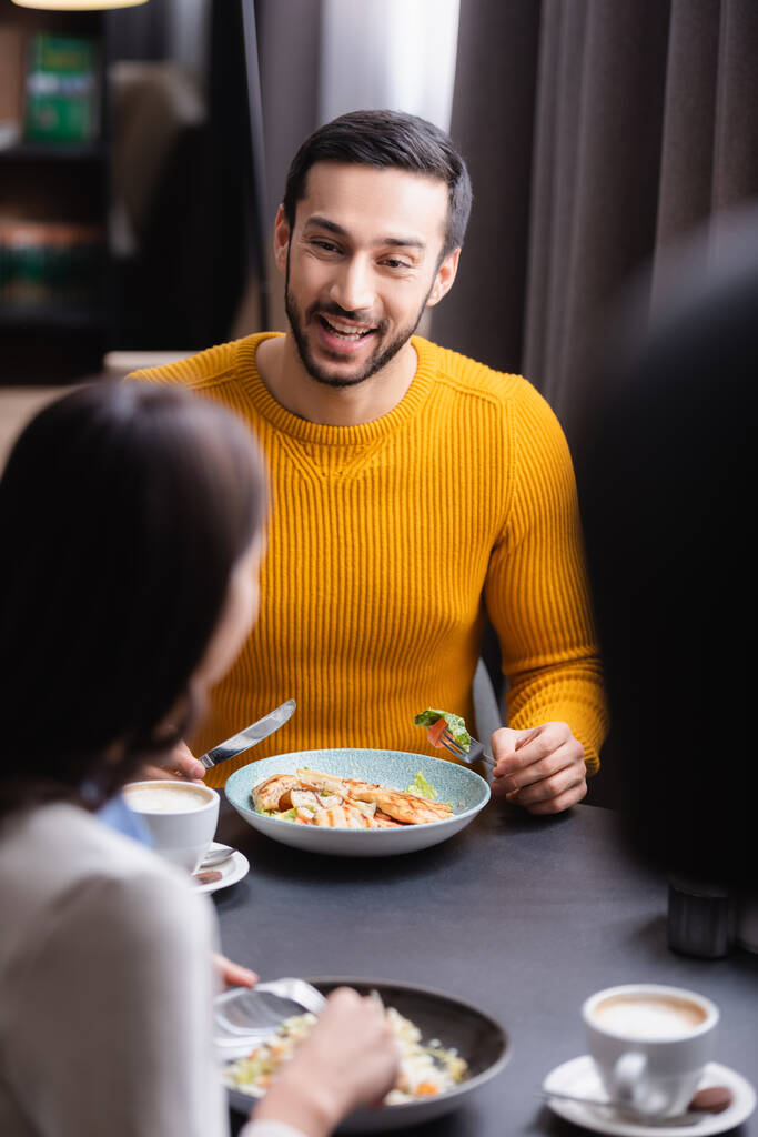Veselý arabský muž drží příbory v blízkosti lahodného salátu, cappuccino a přítelkyně na rozmazané popředí v restauraci  - Fotografie, Obrázek