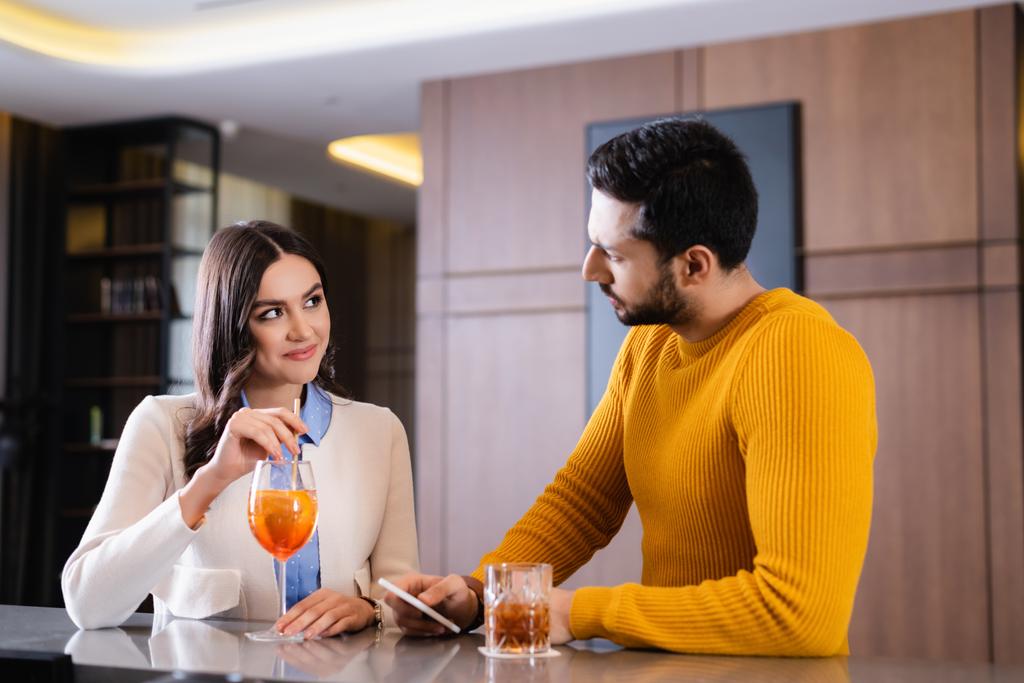 Lächelnde Frau mit Cocktail blickt muslimischen Freund mit Smartphone in der Nähe der Theke an  - Foto, Bild