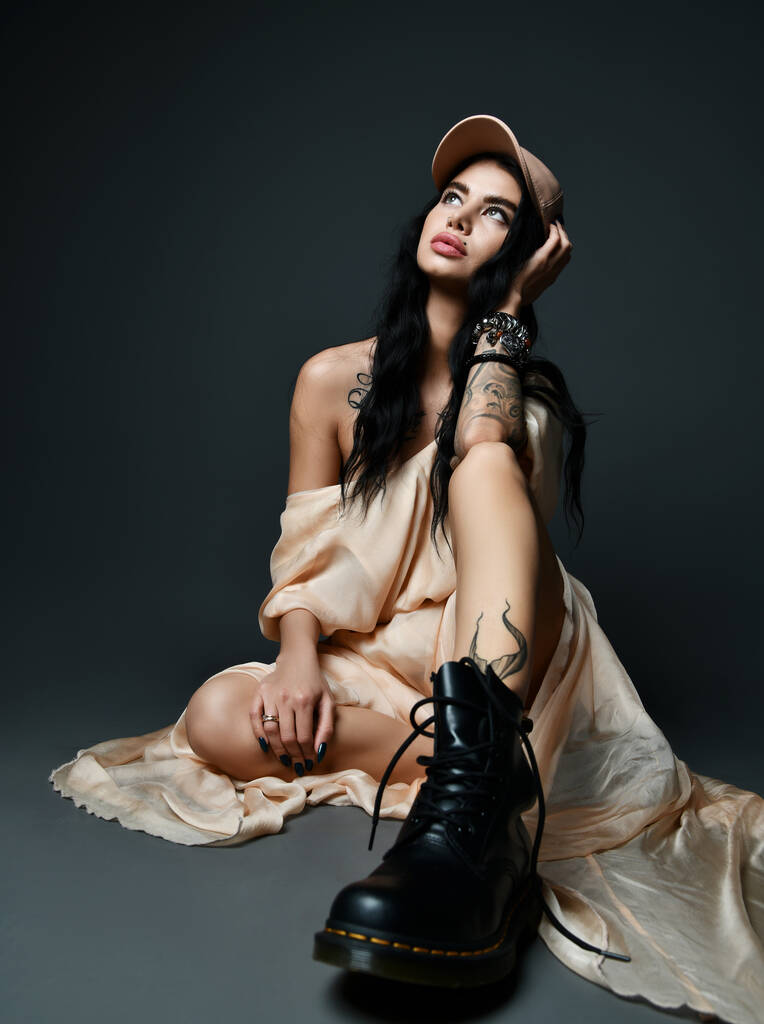 Mulher morena sexy com tatuagem no braço e piercing no rosto no vestido e sapatos brutais maciços senta-se no chão olhando para cima  - Foto, Imagem