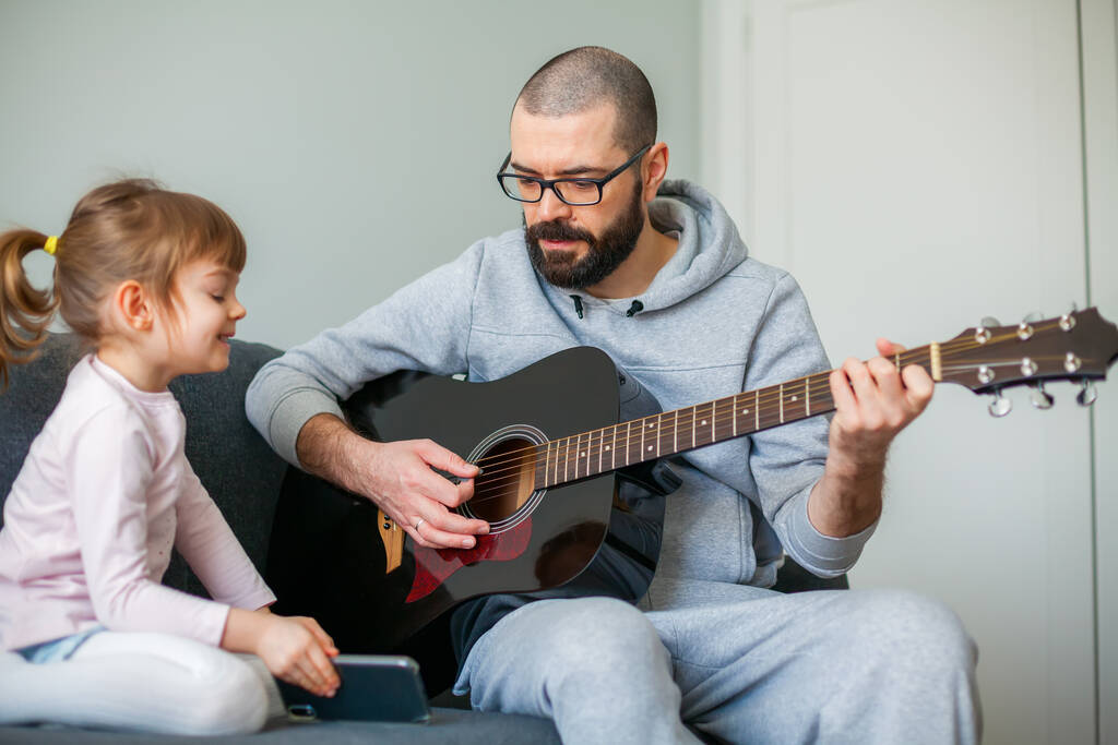 彼女のためにギターを弾いている間、彼は彼女のためにギターを弾いている間、彼女の父に電話で歌を示す女の子 - 写真・画像