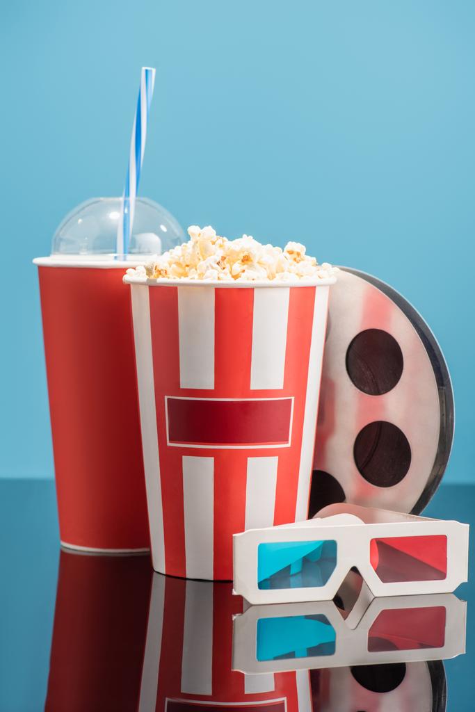 Κύπελλο σόδα, κουβάς με ποπ κορν, 3d ποτήρια και ταινία bobbin σε γυαλιστερή επιφάνεια που απομονώνονται σε μπλε, κινηματογραφική έννοια - Φωτογραφία, εικόνα