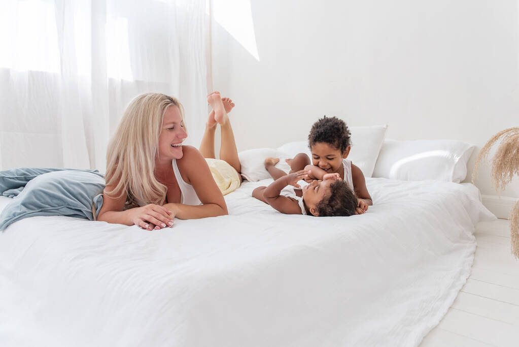 Jeune mère blonde caucasienne joue, se trouve sur un lit blanc avec fille afro-américaine fils. Joyeuse matinée familiale multi ethnique. Femme étreint, caresse les enfants avec tendresse. Intérieur blanc minimaliste - Photo, image