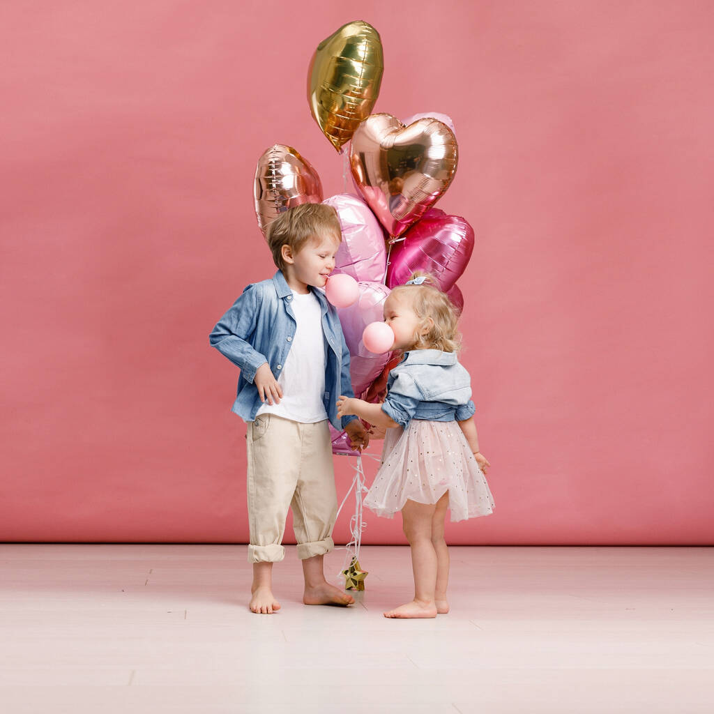 Niño y niña abrazándose, bailando, sonriendo y divirtiéndose cerca de una gran rama de globos rosados en forma de corazón. Concepto de San Valentín. Aislado sobre fondo rosa. - Foto, imagen