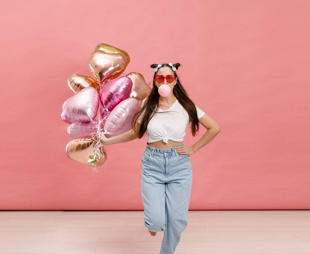 Młoda kobieta tańczy, uśmiecha się i bawi gumą do żucia w pobliżu dużej gałęzi różowych balonów w kształcie serca. Izolacja na różowym tle. - Zdjęcie, obraz