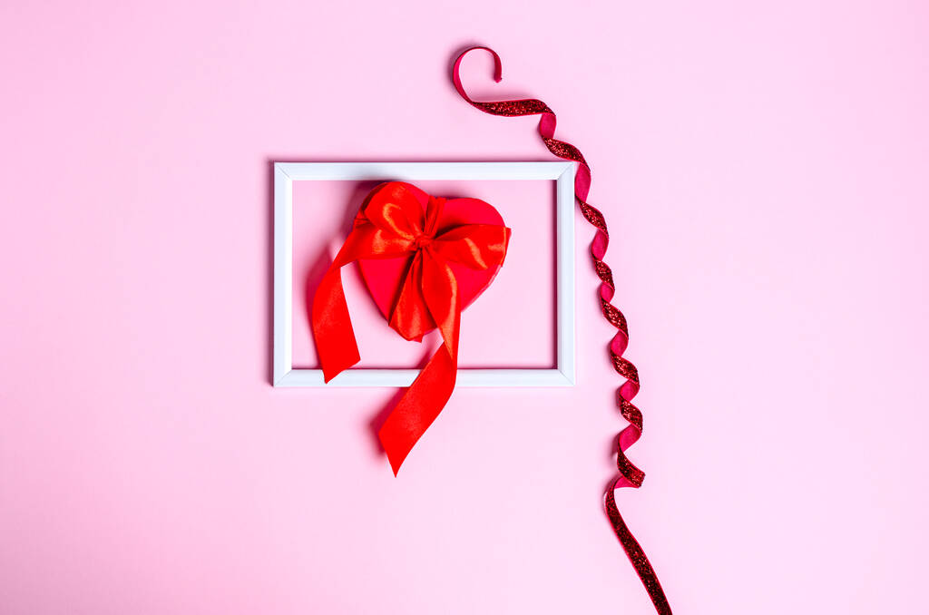 Red box for a gift with a ribbon in the form of a heart in a white frame on a pink background. Love concept, copy space, flat lay, place for text, banner, top view. - Photo, Image