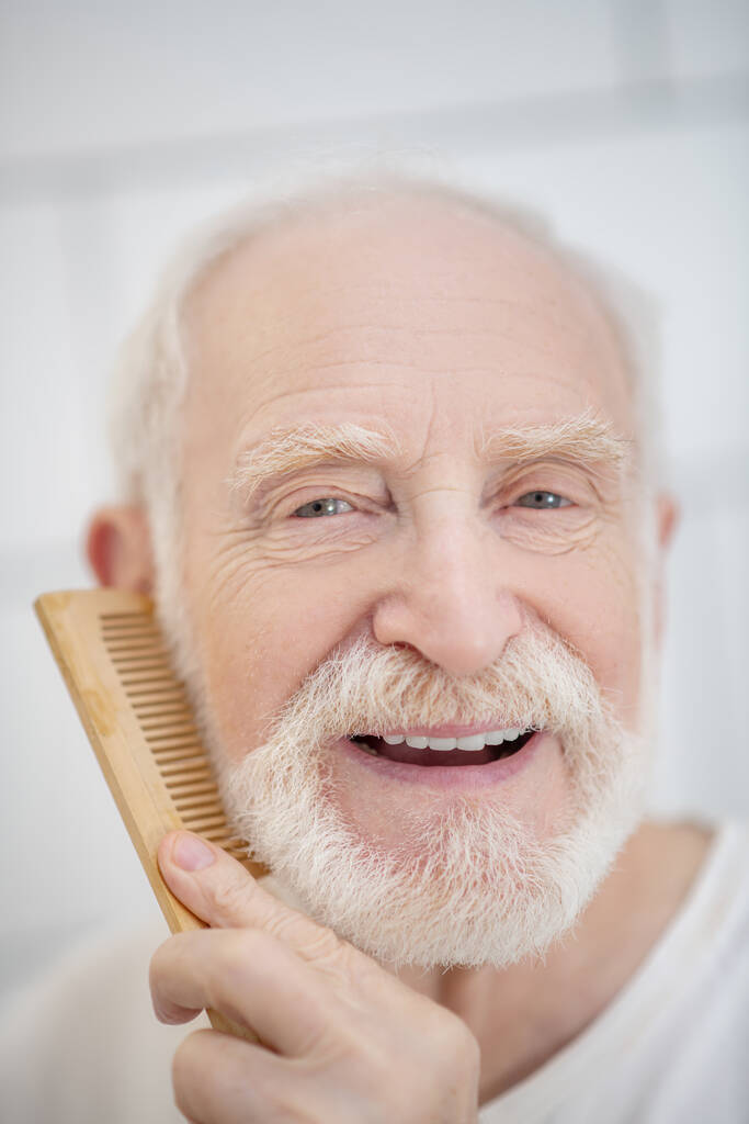 Közelkép egy ősz hajú férfiról, aki a szakállát fésüli. - Fotó, kép