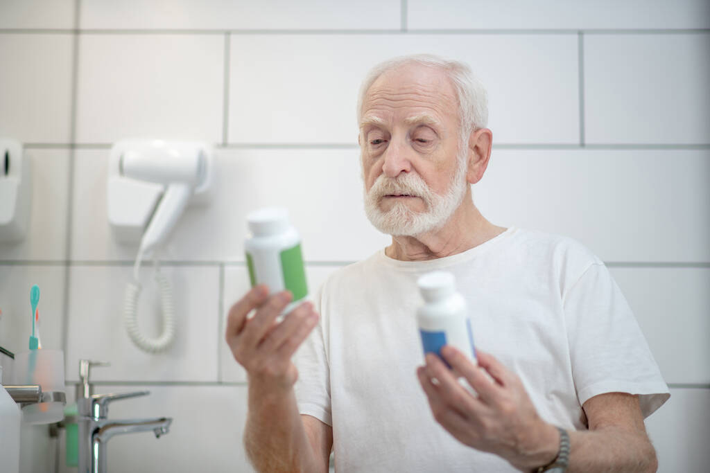 Gray-haired man in white tshirt holding bottles with pills and looking thoughtful - Photo, Image