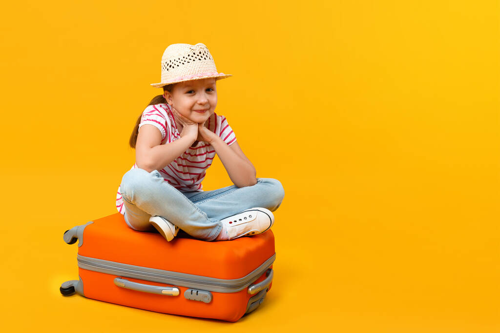 The child sits on a suitcase and holds his cheeks with his palms on a yellow background. Little girl dreams of travel, adventure, vacation. - Photo, Image