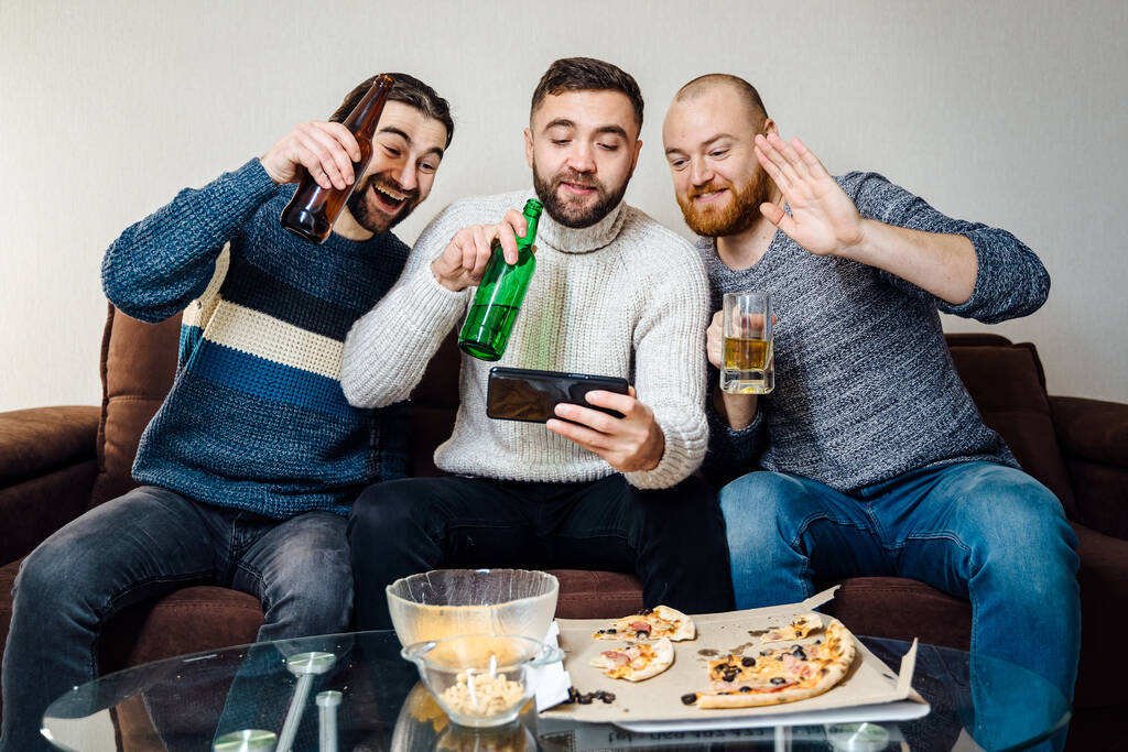 tři veselí přátelé muži zdravit své přátele prostřednictvím video chatu, na telefonu sedí na pohovce doma, pozitivní muži s pivem, on-line diskuse během karantény, izolace doma - Fotografie, Obrázek