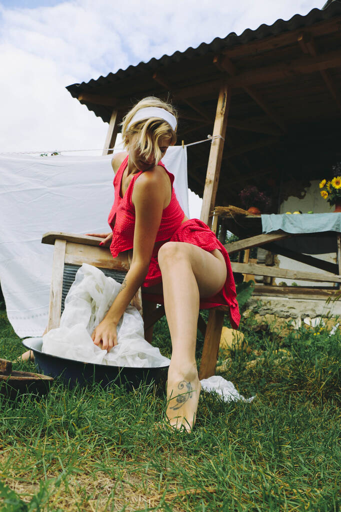 Lavandaria seca. Jovem bela mulher sexy lava roupas e pendura roupas íntimas ao ar livre em um dia ensolarado com um jardim. Em roupas vermelhas. - Foto, Imagem