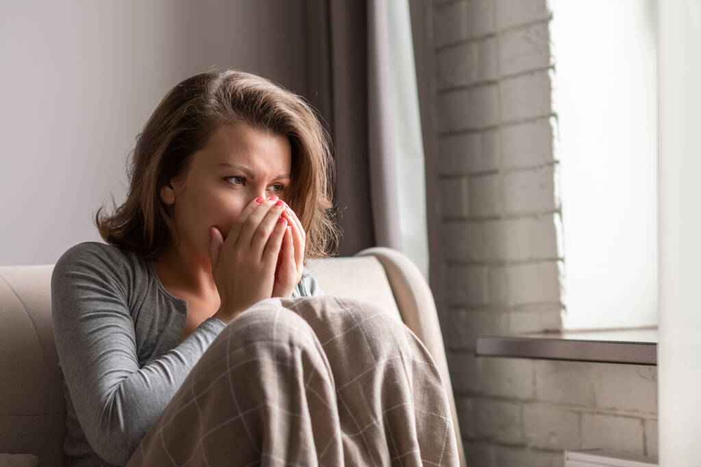 Хвора брюнетка жінка чхає, сидячи на дивані у вітальні біля вікна
 - Фото, зображення