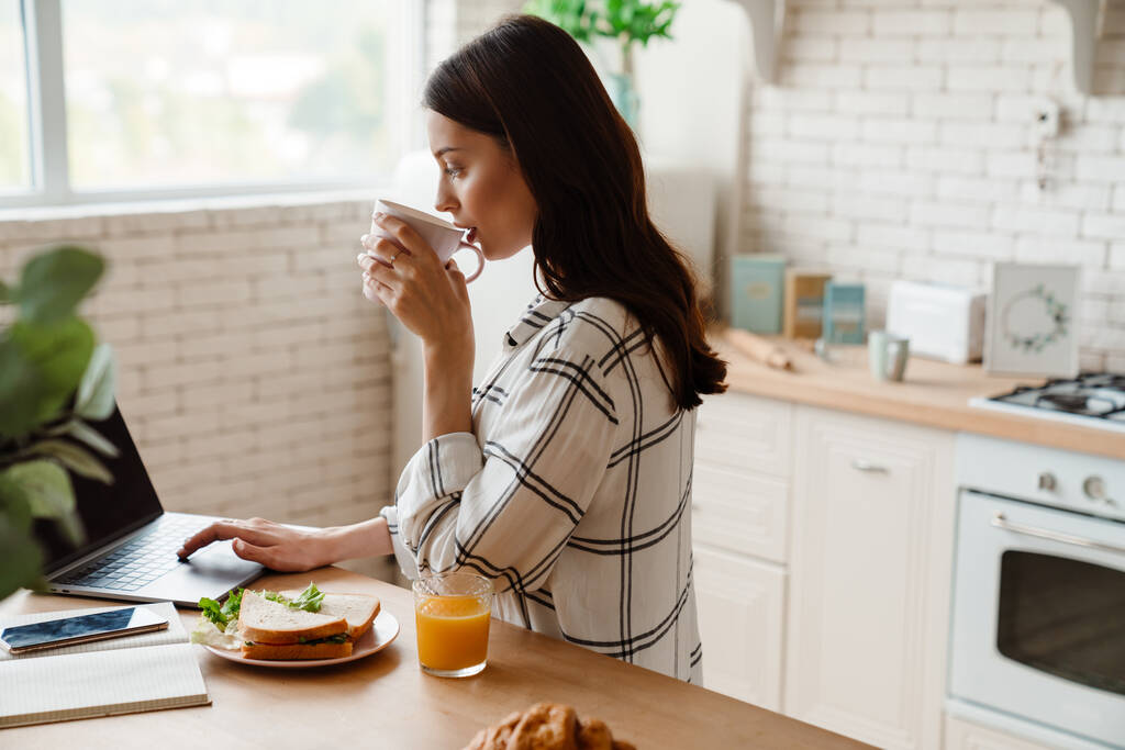 Schöne fokussierte Frau arbeitet mit Laptop beim Frühstück in der gemütlichen Küche - Foto, Bild
