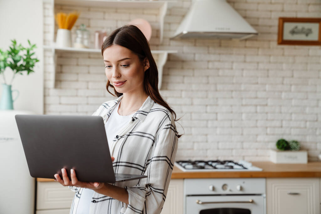 Schöne zufriedene Frau arbeitet mit Laptop, während sie in der gemütlichen Küche steht - Foto, Bild