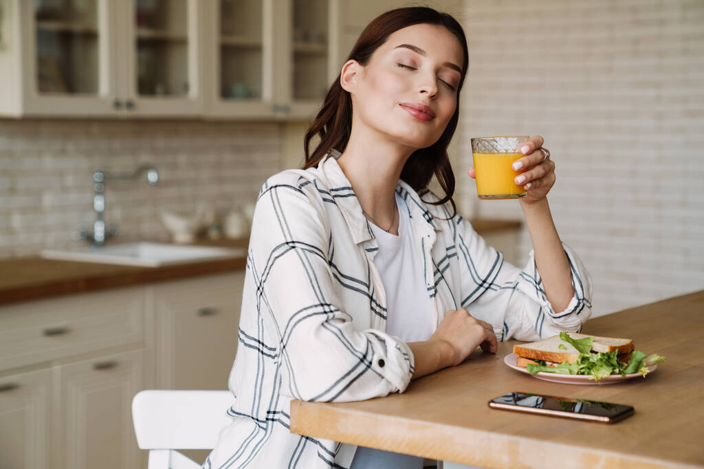 Красива усміхнена жінка п'є сік, снідаючи в затишній кухні
 - Фото, зображення