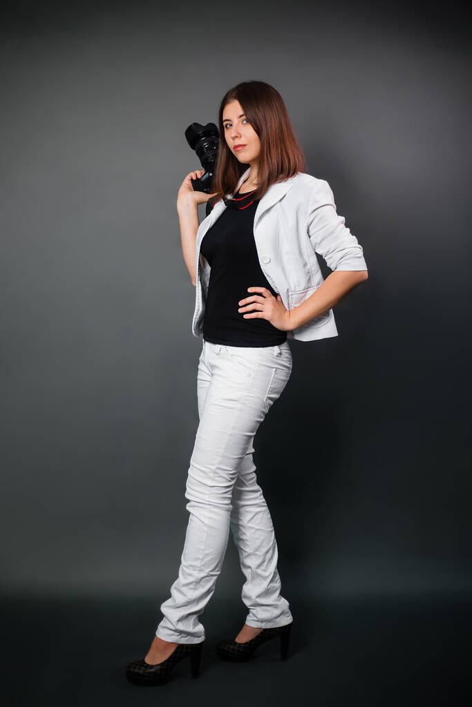 Жінка в білому костюмі з камерою в руках позує, стоячи на чорному тлі. Сучасний фотограф у студії
. - Фото, зображення