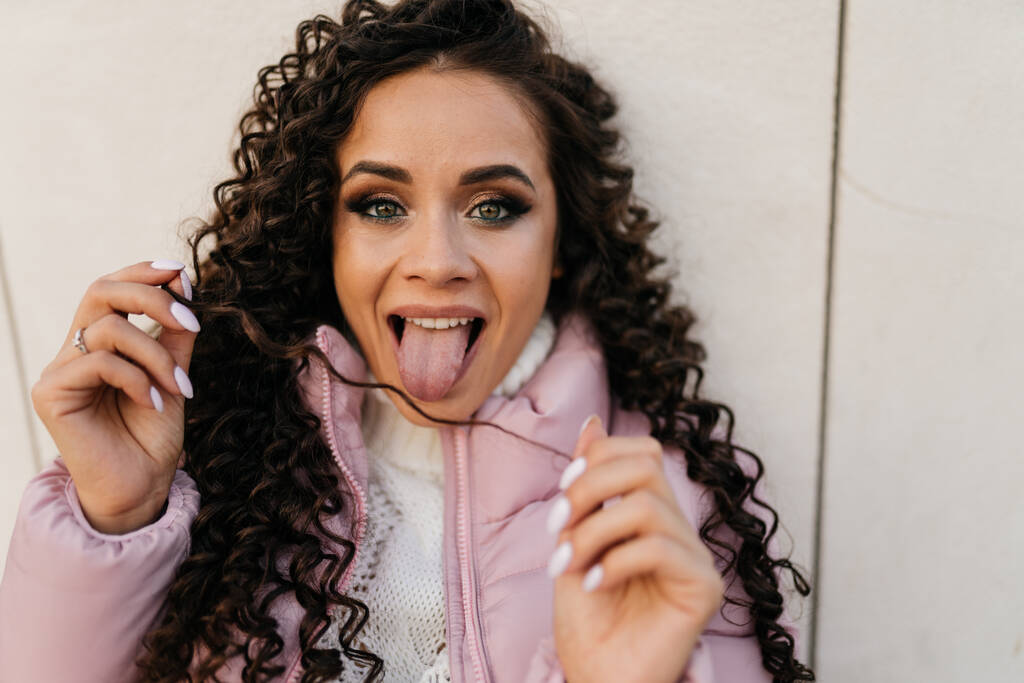 Menina em jaqueta com cabelos longos encaracolados mostra língua para câmera - Foto, Imagem