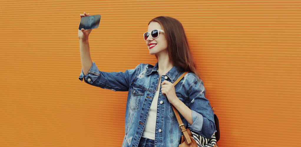 Πορτρέτο του χαμογελαστού γυναίκα λήψη μια φωτογραφία selfie από smartphone φορώντας ένα τζιν σακάκι σε πορτοκαλί φόντο - Φωτογραφία, εικόνα