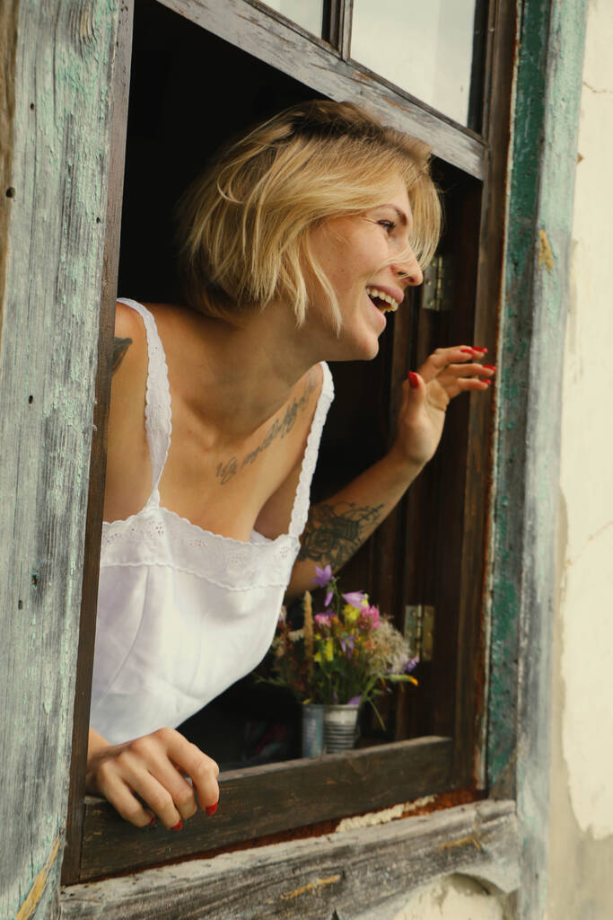 Retrato de arte de una hermosa mujer joven, mira a través de una vieja ventana se asoma, se sienta en el alféizar de la ventana en estilo grunge. - Foto, imagen