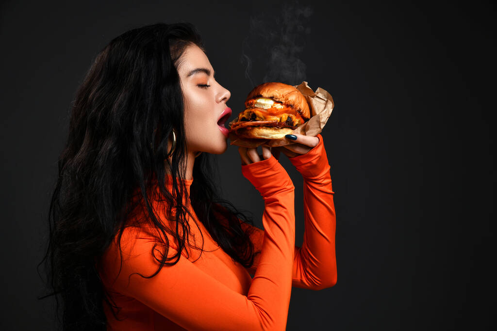 Portret van hete sexy brunette vrouw met pruillip in rode kleren eten bijten grote smakelijke ambachtelijke hamburger met stoom - Foto, afbeelding
