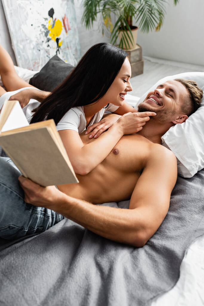Vrolijke vrouw aanraken kin van sexy man met boek op bed  - Foto, afbeelding