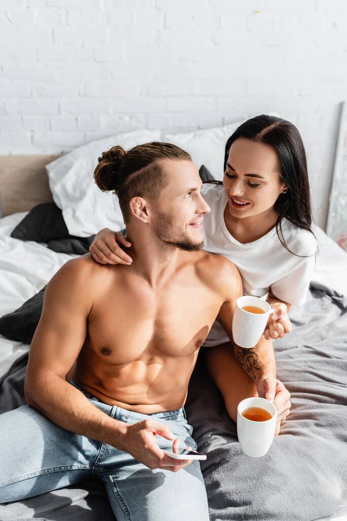 Homme sexy tenant smartphone près femme souriante avec tasse sur le lit  - Photo, image