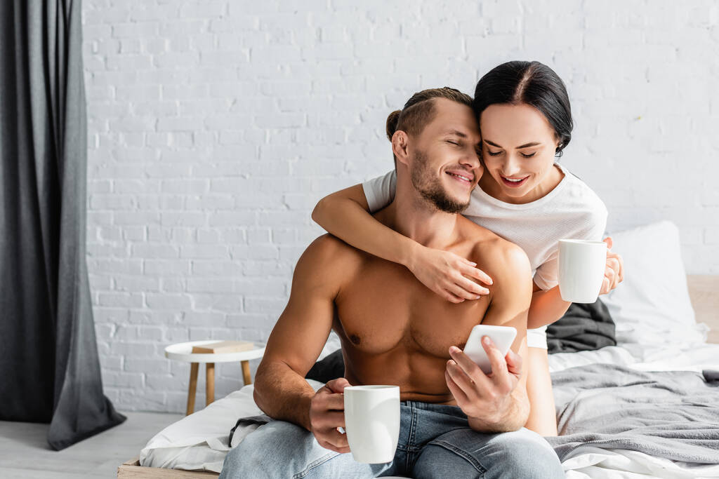Mulher sorridente com copo abraçando homem sem camisa com smartphone na cama  - Foto, Imagem