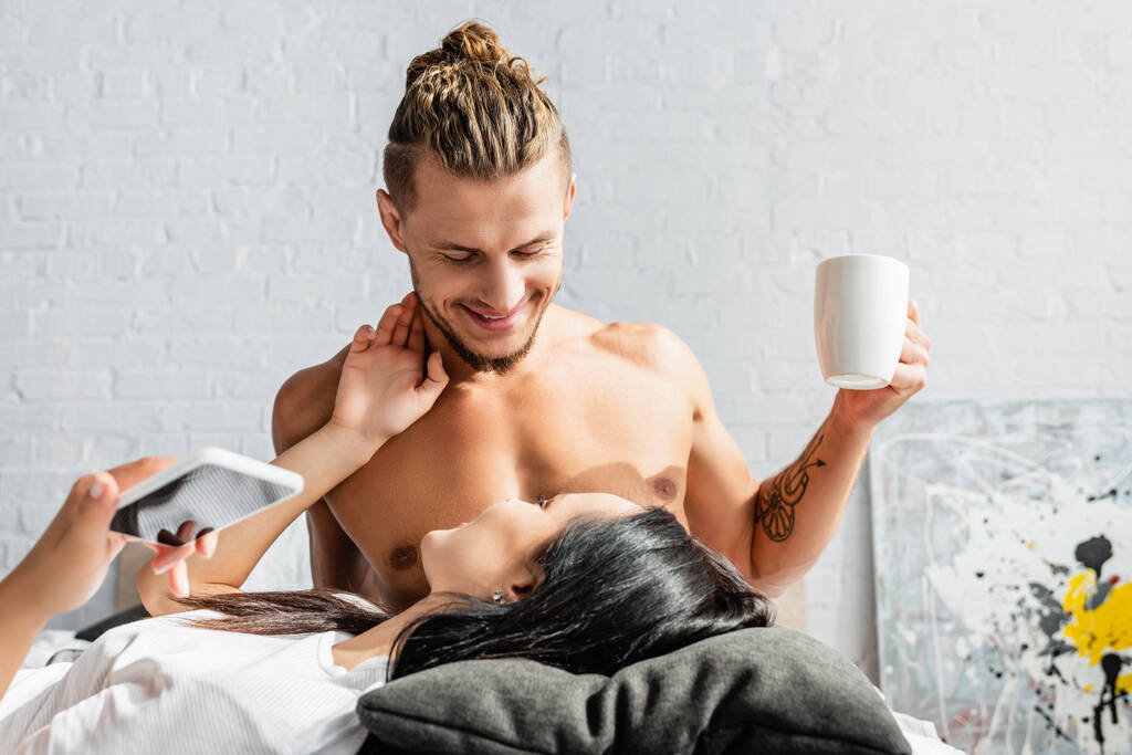 Homme souriant tenant tasse près de petite amie avec smartphone au premier plan flou  - Photo, image