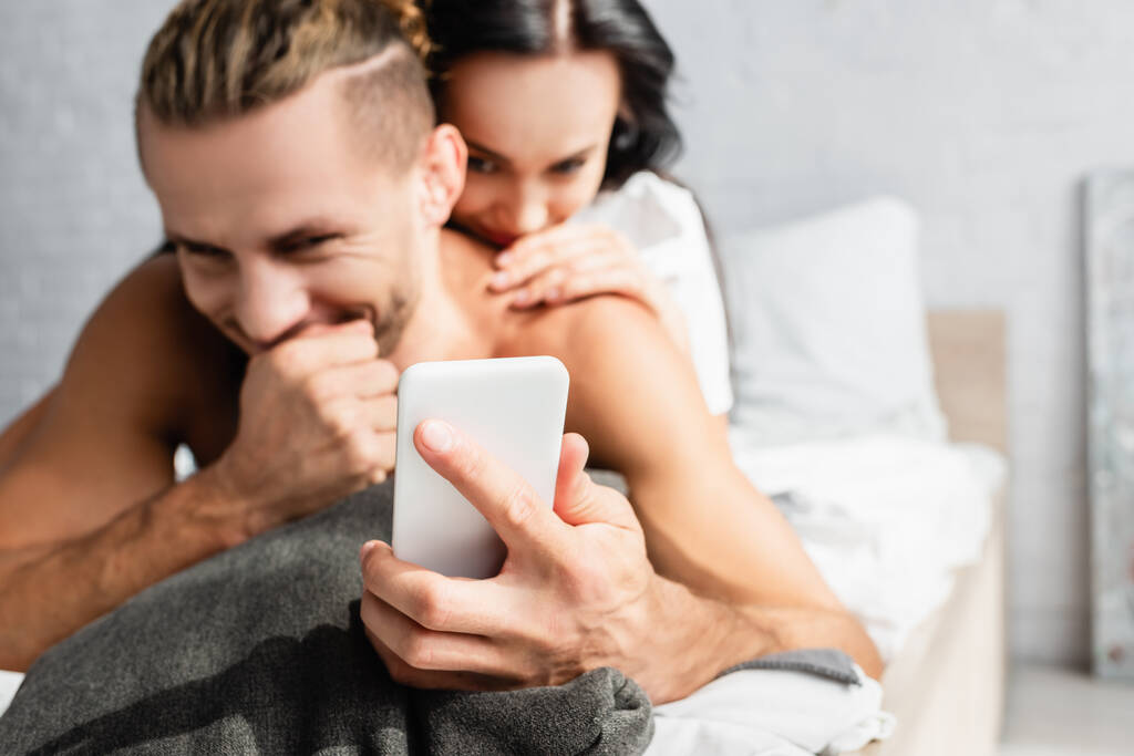Telefon komórkowy w ręku mężczyzny bez koszulki w pobliżu dziewczyny na rozmytym tle w sypialni  - Zdjęcie, obraz