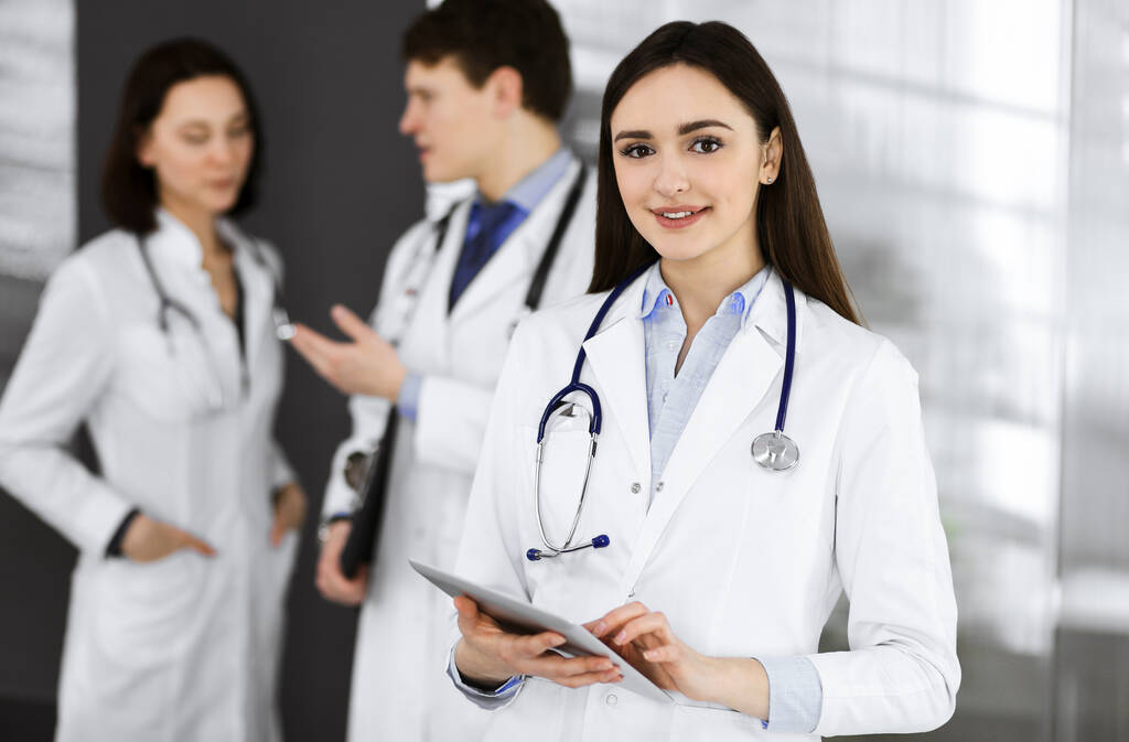 Una doctora inteligente sonriente sostiene una tableta en sus manos, mientras está de pie junto con sus colegas en una clínica. Médicos en el trabajo. Servicio médico perfecto en un hospital - Foto, Imagen