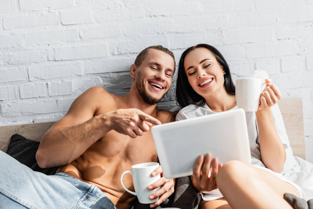 Веселая пара с чашками с помощью цифрового планшета на размытом переднем плане на кровати  - Фото, изображение