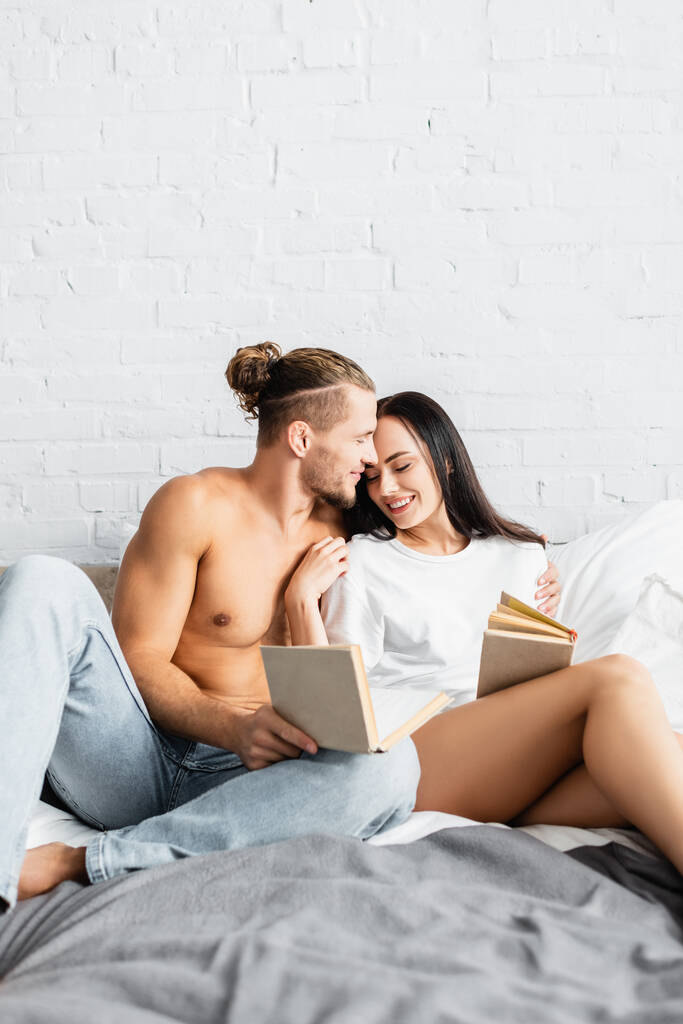 Sorrindo homem sem camisa abraçando namorada com livro na cama  - Foto, Imagem