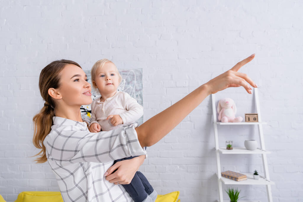 sonriente mujer apuntando con el dedo mientras sostiene a su hijo pequeño - Foto, imagen