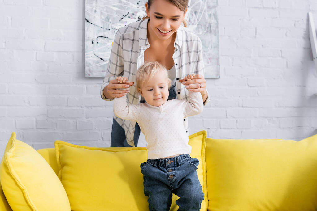fröhliche junge Frau hält Hände eines kleinen Jungen auf gelbem Sofa - Foto, Bild