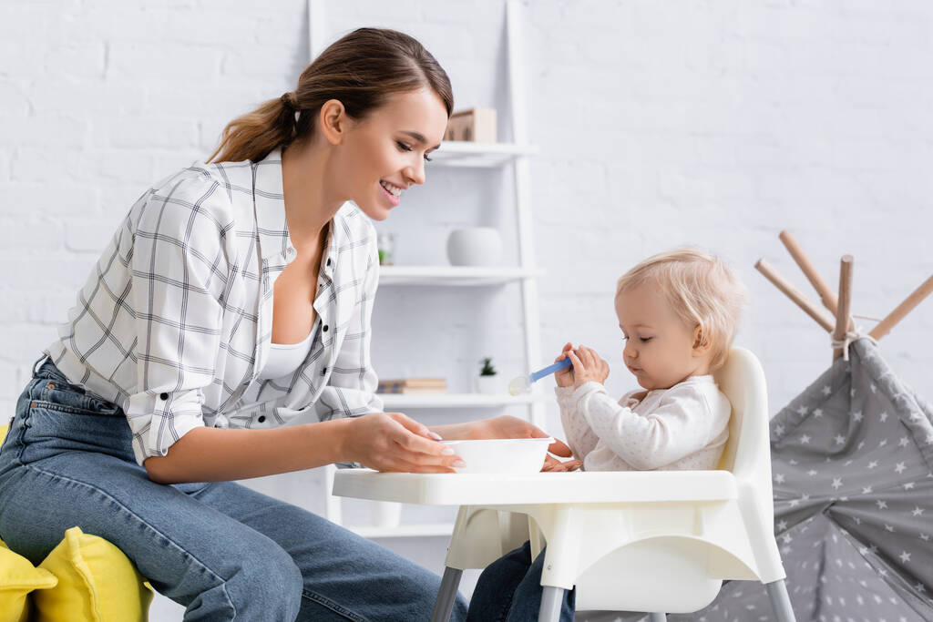 little boy sitting on kids chair with spoon near happy mother - Photo, Image