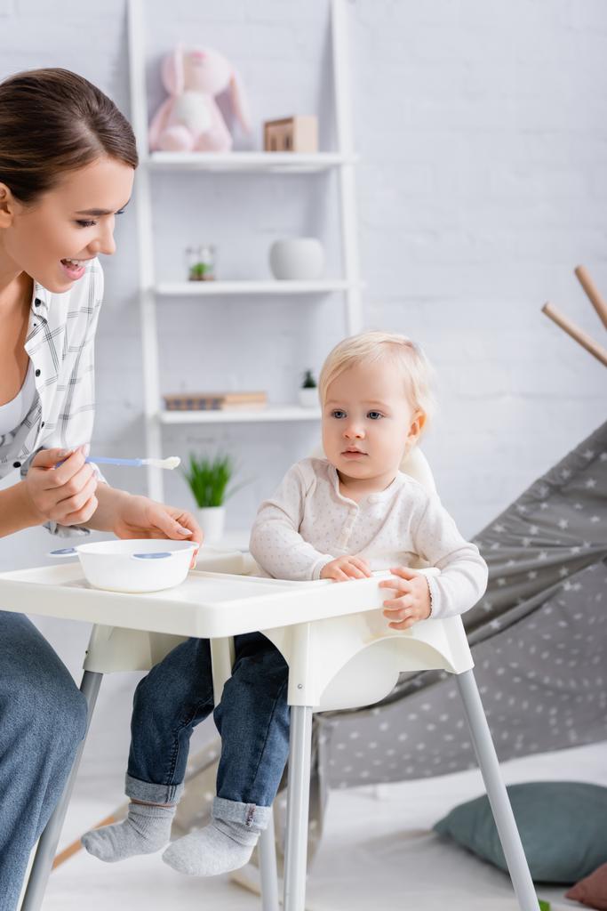 young woman holding spoon with puree near child sitting on kids chair - Photo, Image