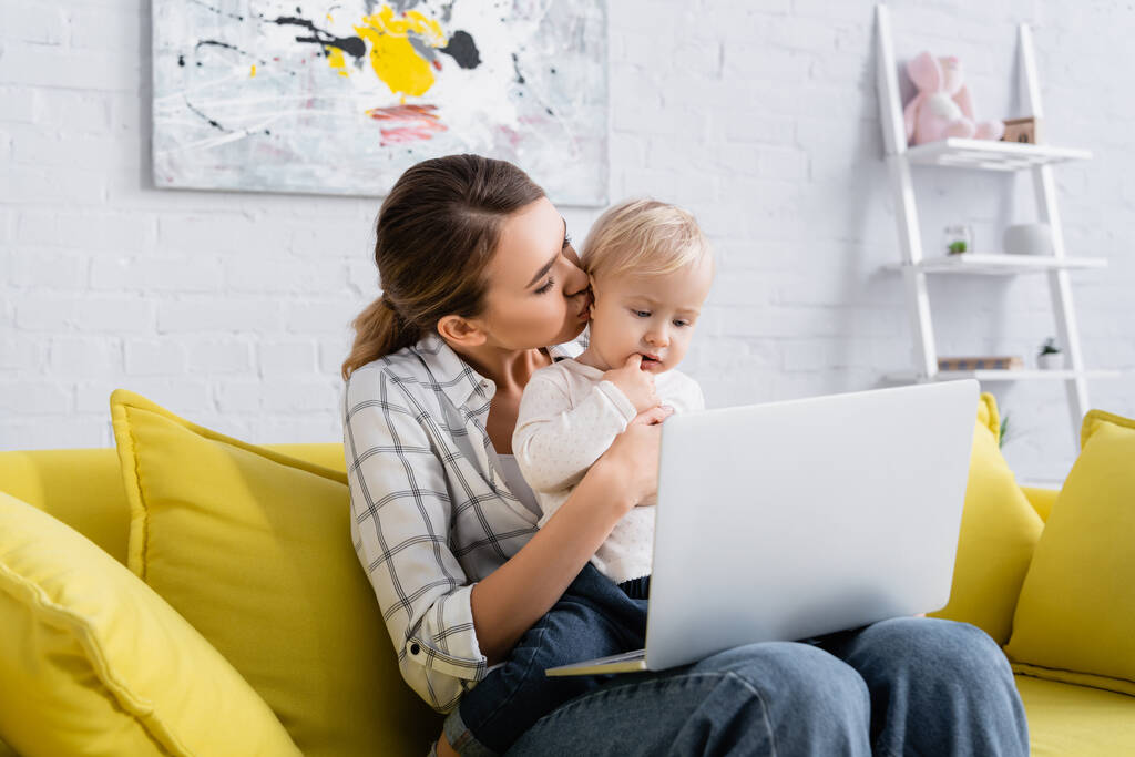 jeune freelance embrasser petit enfant tout en étant assis sur le canapé jaune avec ordinateur portable - Photo, image