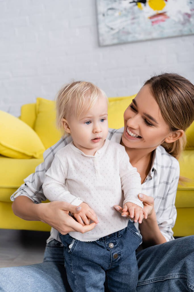 alegre mujer mirando bebé niño mientras sosteniendo sus manos en casa - Foto, Imagen