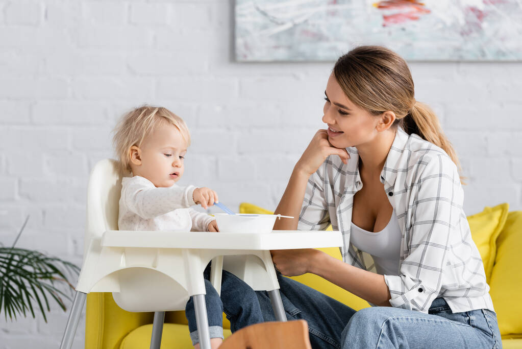 feliz madre mirando hijo sentado en niños silla con cuchara - Foto, Imagen