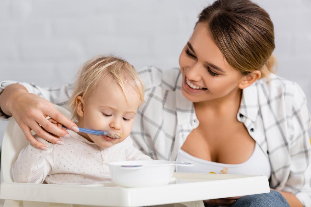 junge lächelnde Frau füttert Sohn auf Kinderstuhl - Foto, Bild
