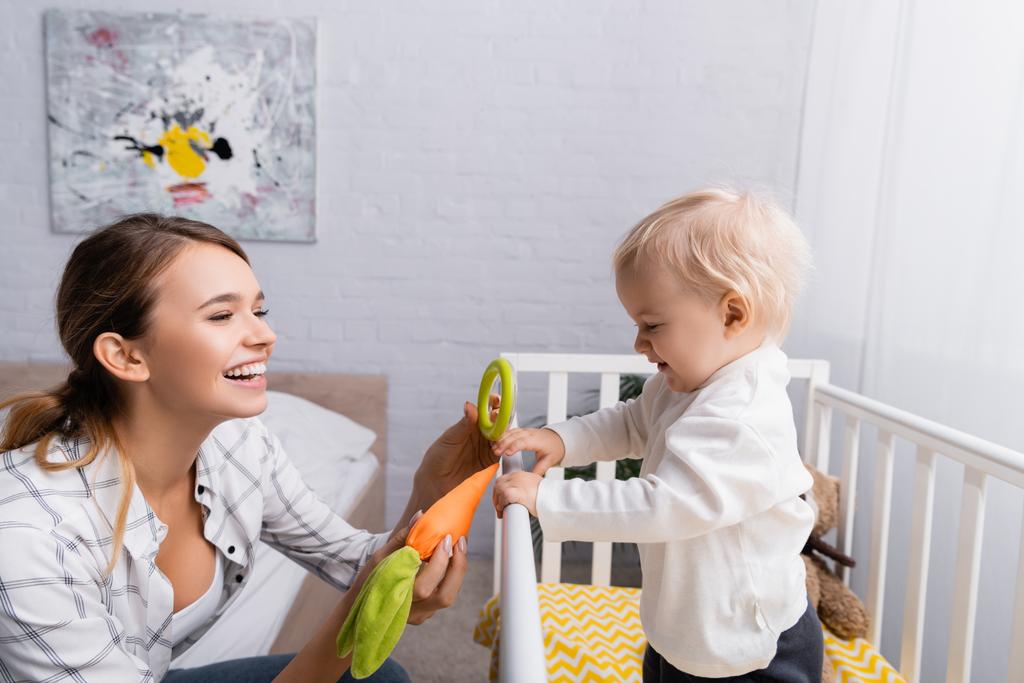 cheerful woman holding toy carrot and rattle near toddler boy standing in crib - Photo, Image