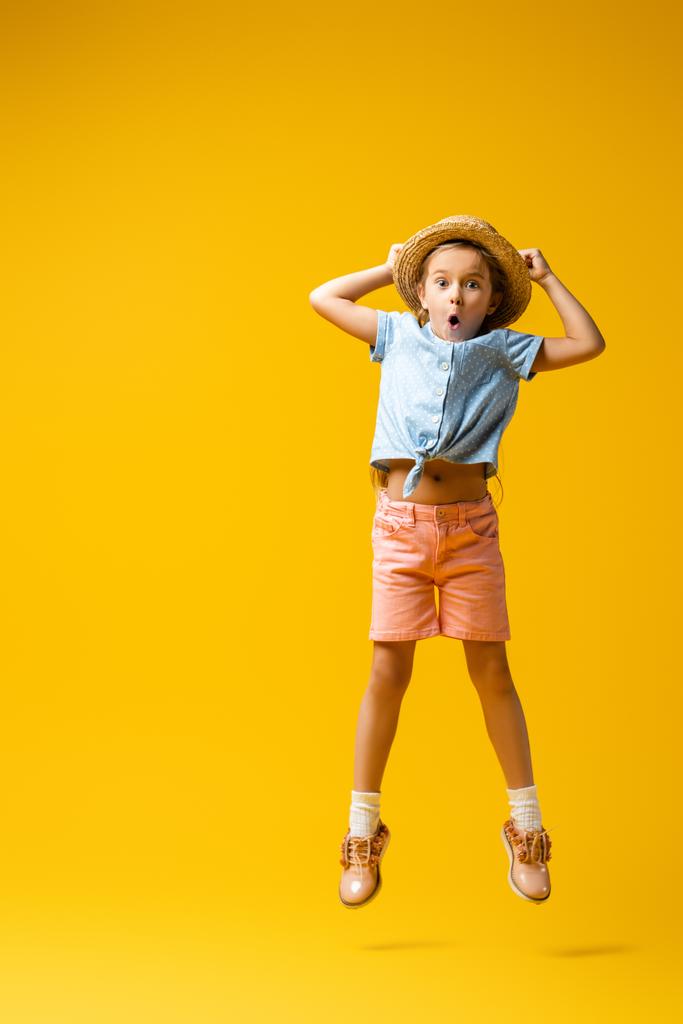 longitud completa de niño sorprendido en sombrero de paja levitando en amarillo - Foto, imagen